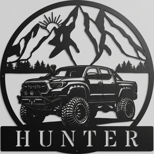 Custom offroad Toyota truck metal sign in powder-coated matte black, featuring mountain peak backdrop and personalized name plate, mounted on a light gray studio wall.